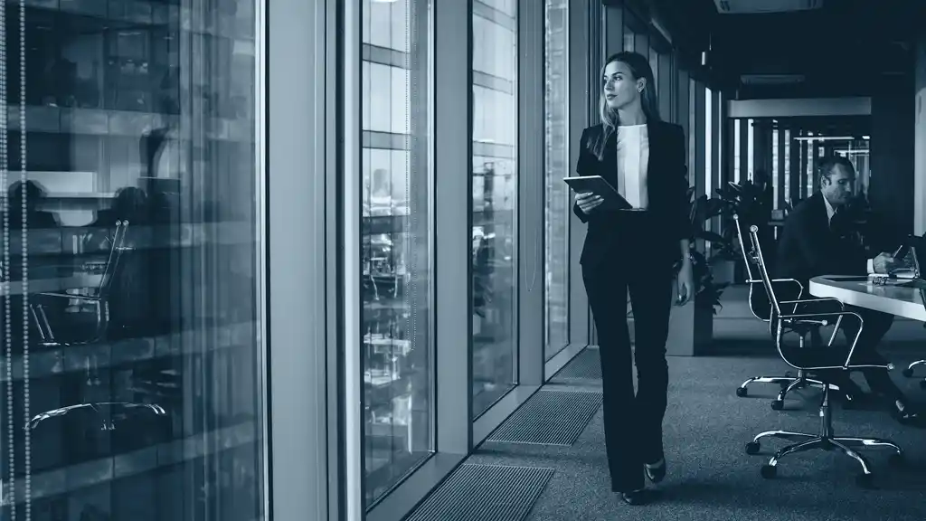 Serious Female Administrative Manager wearing Formal Suit Walking In Office while gazing out wall of windows
